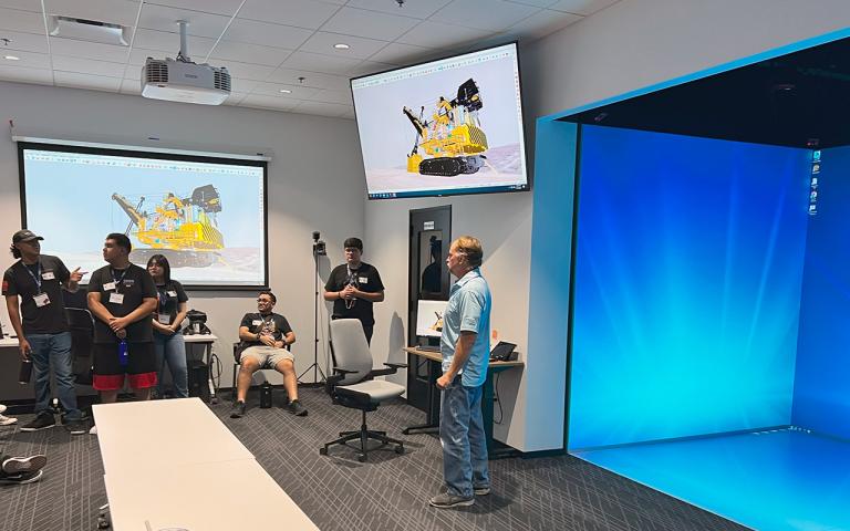 Students listen to a man in a light blue shirt talk about the wall which has images projected onto it.