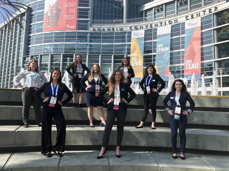 A group of women stand spaced out on a set of steps in front of a large glass building.