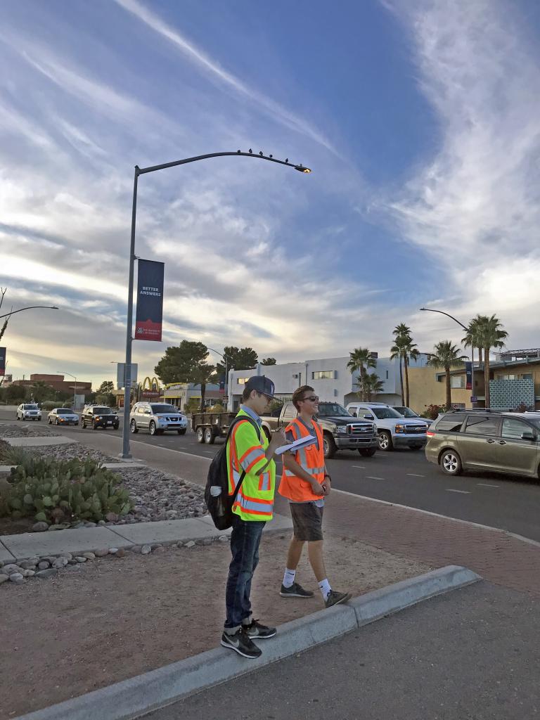 Two students wearing bright orange and yellow vests stand at an intersection along Speedway as the sun sets behind them.