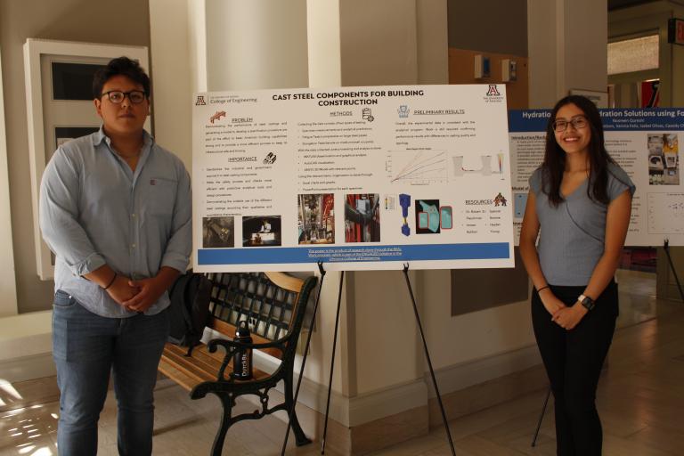 A young man and a young woman stand on either side of a scientific poster that reads, "Cast Steel Components for Building Construction."