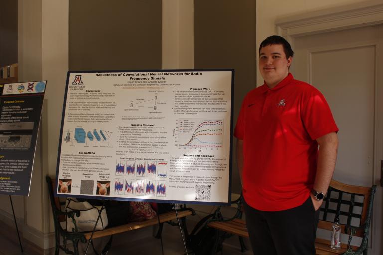 A young man stands next to a scientific poster that reads, "Robustness of Convolutional Neural Networks for Radio Frequency Signals"