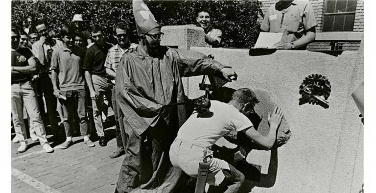 A man wearing a robe and a hat with a three-leaf clover on it whacks another young man on the back while the second man leans down to kiss a stone embedded in a concrete slab.