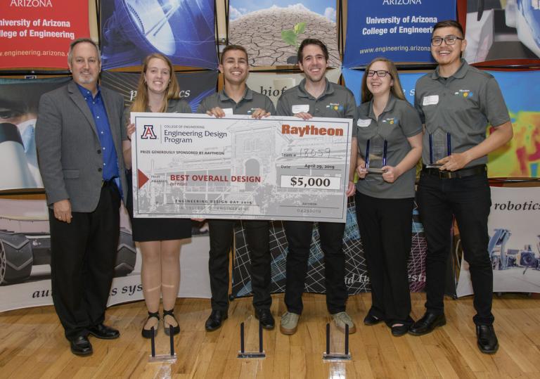 A group of five students in matching grey polos and a man in a blazer hold up a giant, $5,000 check, made out to "Best Overall Design."