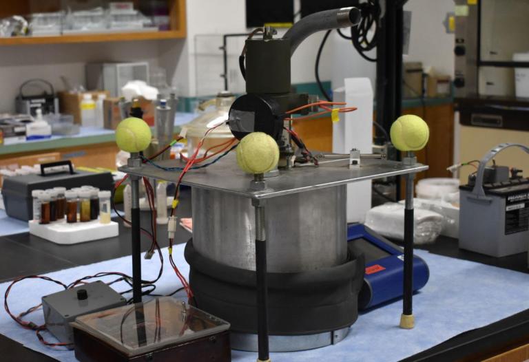 A metal cylinder with wiring attached to it and four tennis balls on the corners of a square support frame.