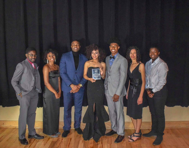 A group of seven black students in formal clothing smile for a photo. The woman in the center holds a plaque.