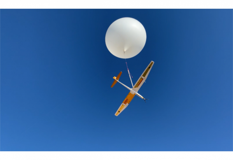 an orange and white sailplane attached to a large white balloon, against a blue sky.