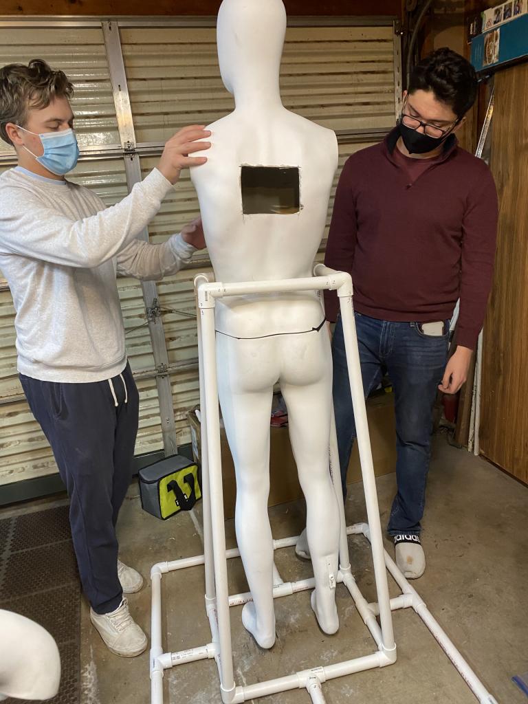Two students wearing face masks attach arms to a white mannequin, which has a hole carved in its back and is held upright by a PVC pipe base.