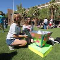 A woman with blond hair and a woman with brown hair add tape to their handmade oven on a grassy lawn.