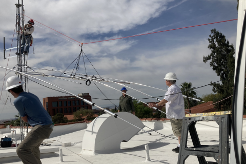 Five people on a roof installing an antenna and wearing hard hats. Four are on the surface of the roof while one is climbing up a metal structure.