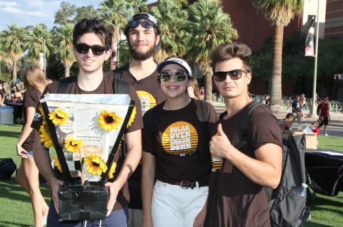Four students gather around their solar oven, which is made of cardboard and foil and decorated with plastic sunflowers