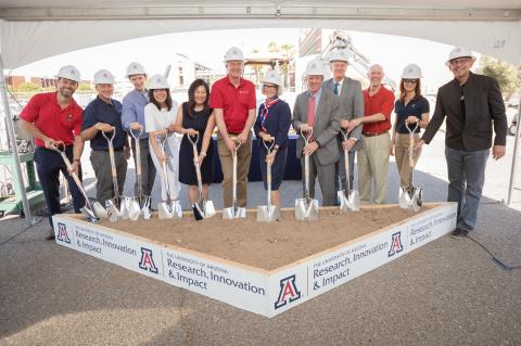A group of a dozen people wearing white construction hats hold shovels that are placed into a square of dirt.