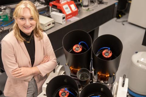 A young woman wearing a blazer is crossing her arms and smiling. She's standing in a lab environment, and next to her are four black cylinders, approximately 10 inches in diameter, win imaging instrumentation inside.