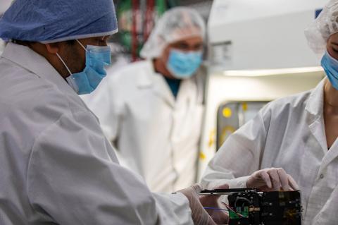 Two masked students work in a cleanroom with satellite components