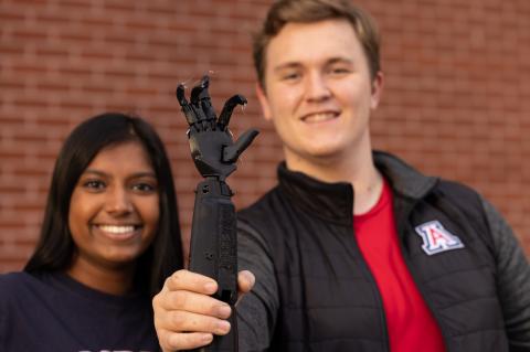 Nisha Rajakrishna and Collin Preszler in the background. Collin is holding up a black prosthetic arm in the foreground.