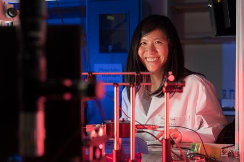 Judith Su wearing a white labcoat and smiling behind a complicated array of sensors.