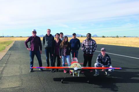 A group of eight people stand smiling for a photo with a hand-built airplane, which sits in front of them and looks to have a wingspan of about 10 feet.