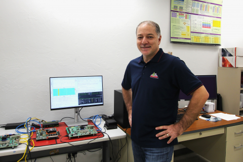 Ali Akoglu in his lab. His hands are on his hips and he is smiling. Behind him is a computer monitor and a green circuit board.