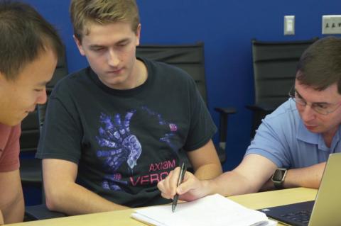 Three men lean over a sheet of paper and a laptop, the one on the far right pointing and explaining a concept.