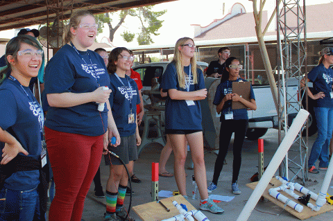 A group of high school girls wearing matching blue "Summer Engineering Academy" t-shirts grin off into the distance at a potato (off frame) they just launched out of a homemade potato launcher.