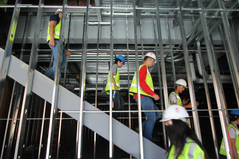 A group of students in yellow vests and hard hats walk down a set of stairs behind a set of beams