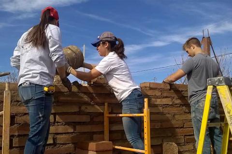 Three students wearing t-shirts and jeans stand on ladders to build a brick wall.