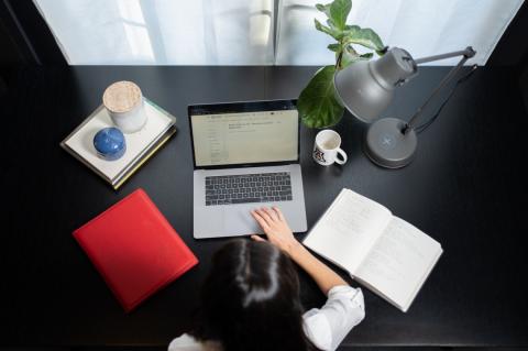A photo of a woman at a desk, taken from overhead. the desk has a laptop, an open notebook, a stack of folders, a plant, a lamp and a University of Arizona coffee mug.