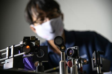 A set of optical devices in the foreground, with DK Kang in the background, blurred and wearing a white face mask.