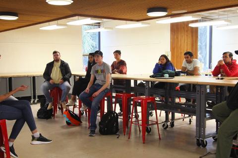 One student addresses a group of others in a room with tables and stools.