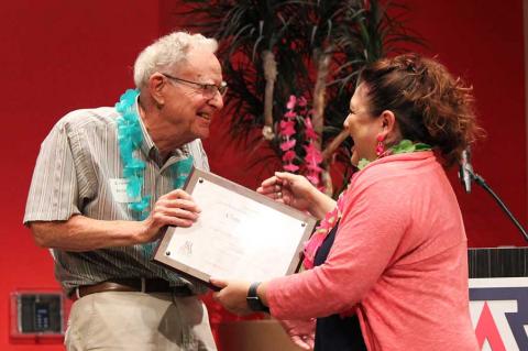 William Cosart, wearing a blue lei, hands a framed certificate to Claudia Arias.