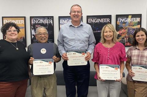 Five people stand indoors with certificates