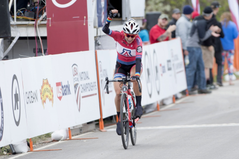 Erica Clevenger, on her bicycle wearing a UA jersey, raises her fist above her head in victory