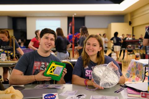 Two students sit at a booth for the Engineering Club showcase and smile. He is holding a green and yellow box and she is holding a circular metallic object..