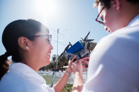 two students, seen from behind, examine a drone. the sun is shining and they are smiling.