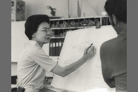 Frances Walker explaining a blueprint on a large white sheet of paper to an onlooker.