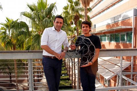 Two men stand in front of a railing and some palm trees, holding a drone encased in a spherical cage, about three feet in diameter