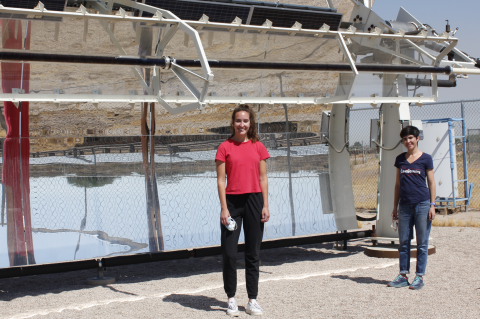 Two women stand in front of a solar-powered desalination system, shaped like a giant curved mirror
