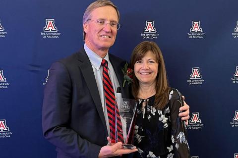 Mike and Sheri Hummel in front of a UA backdrop with a glass award