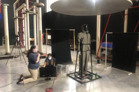 A mannequin covered in black tape is held up by a PVC pipe frame in a large room. Two students kneel next to electronic equipment near the mannequin.