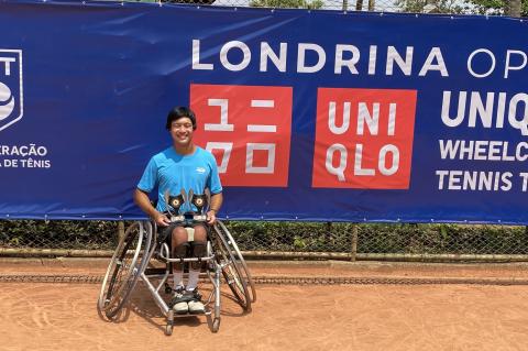Jason Keatseangsilp sits in a sports wheelchair with slanted wheels, smiling and holding two trophies on his lap. Behind him is a blue banner that reads, "Londrina Open UNIQLO Wheelchair Tennis Tour"
