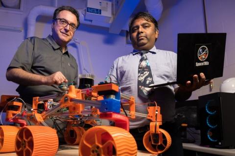 In the foreground, a robot with four orange wheels. Two men stand behind the robot.