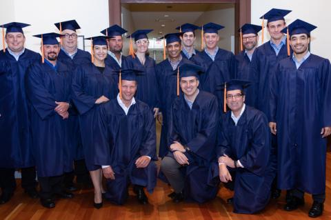 A group of 15 men and women wearing blue graduation caps and gowns