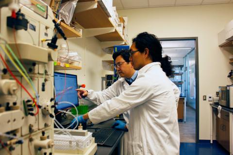 Two researchers in white lab coats look at a computer screen.