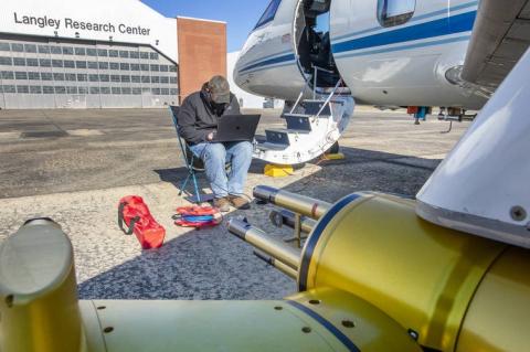 A man sits on a folding chair using a laptop, next to the steps of an airplane. The building behind him has a sign that says, "Langley Research Center."