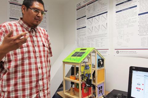 A student stands in front of a house model outfitted with electronic sensors and hooked to a monitor