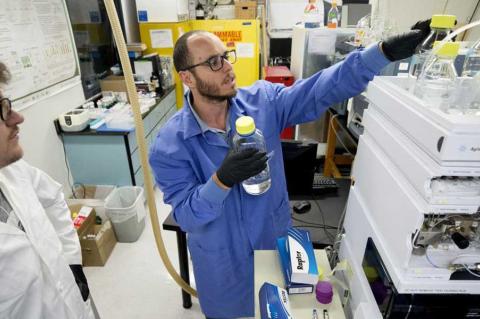 Two men in a laboratory reach for water samples setting on top of a machine.