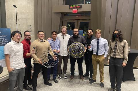 Nine men stand in a row. Two are holding large, spherical mesh robots.