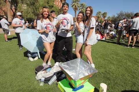 Four students in white shirts smile and pose for a photo on a grassy lawn with an oven made of aluminum foil and cardboard at their feet.