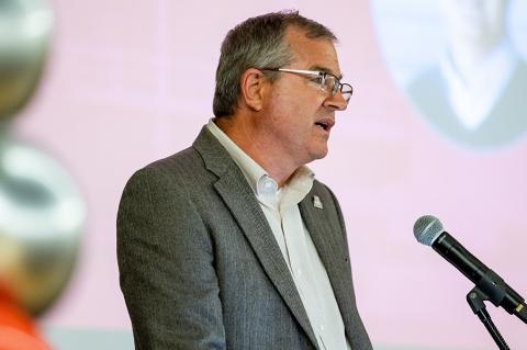 A man in a gray suit gives a speech to an audience of around 150 people.