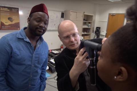 Wolfgang Fink uses an optical device to examine a young woman's eyes, while another young man looks on in the background.