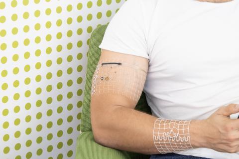 A photo of a man's torso, sitting and holding a cellphone. He has mesh bands with embedded electronics wrapped around his bicep and forearm.